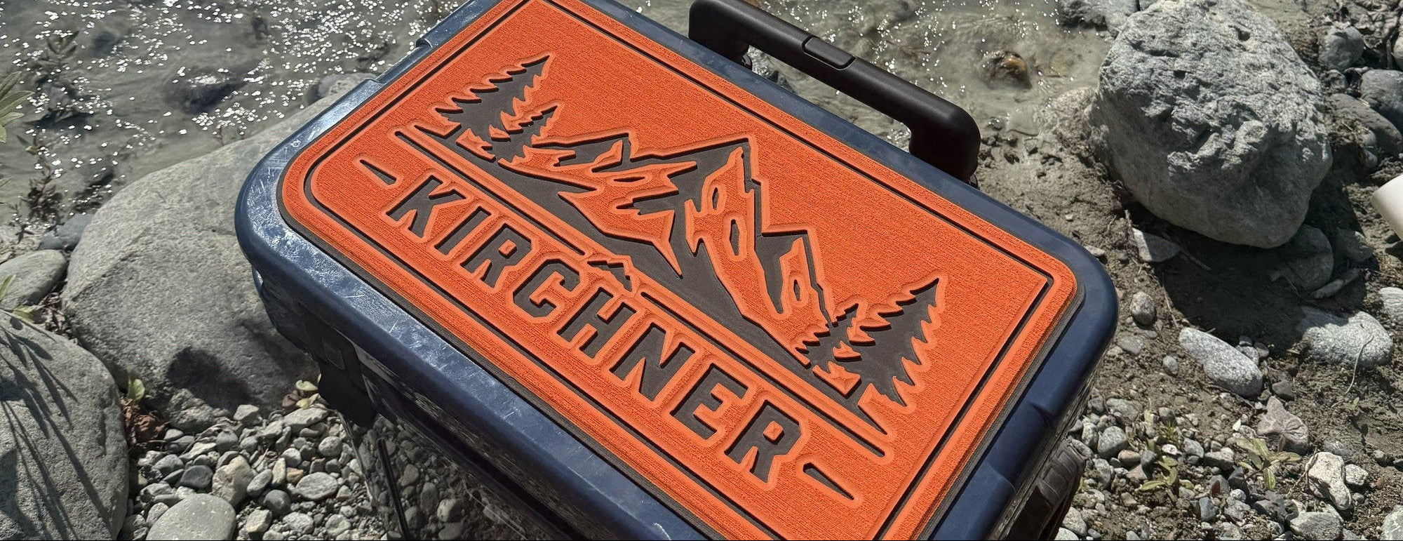 Custom name cooler top pad with mountain scene on a cooler beside a river